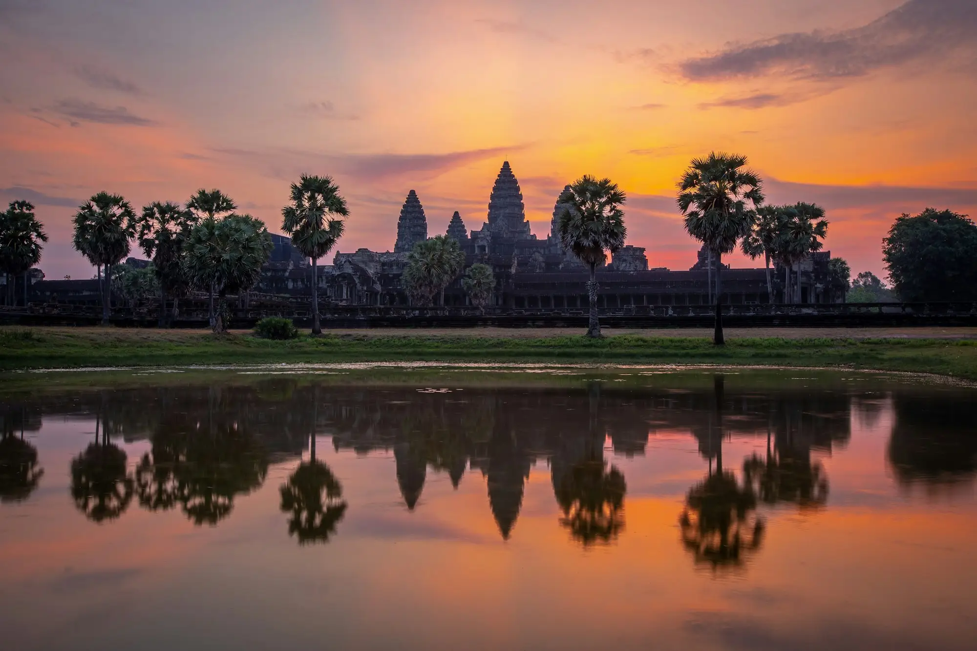 Silhouette of Angkor Wat and palm trees at sunrise with the sky reflected in the pond